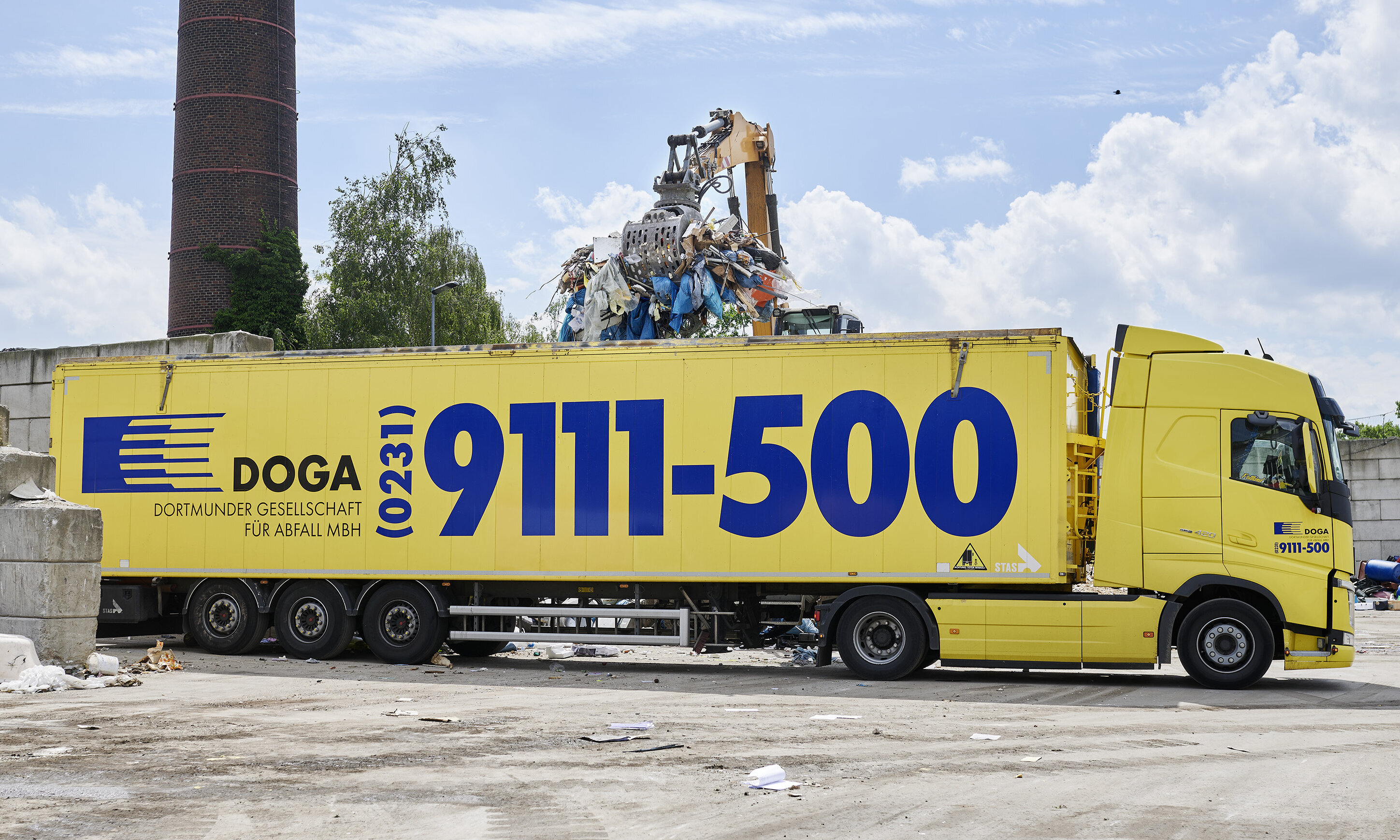 Gelber Lkw mit der Aufschrift ‚DOGA‘ und ‚9111-500‘ steht auf einem Industriegelände; auf der Ladefläche befinden sich Schrott und Maschinenteile.